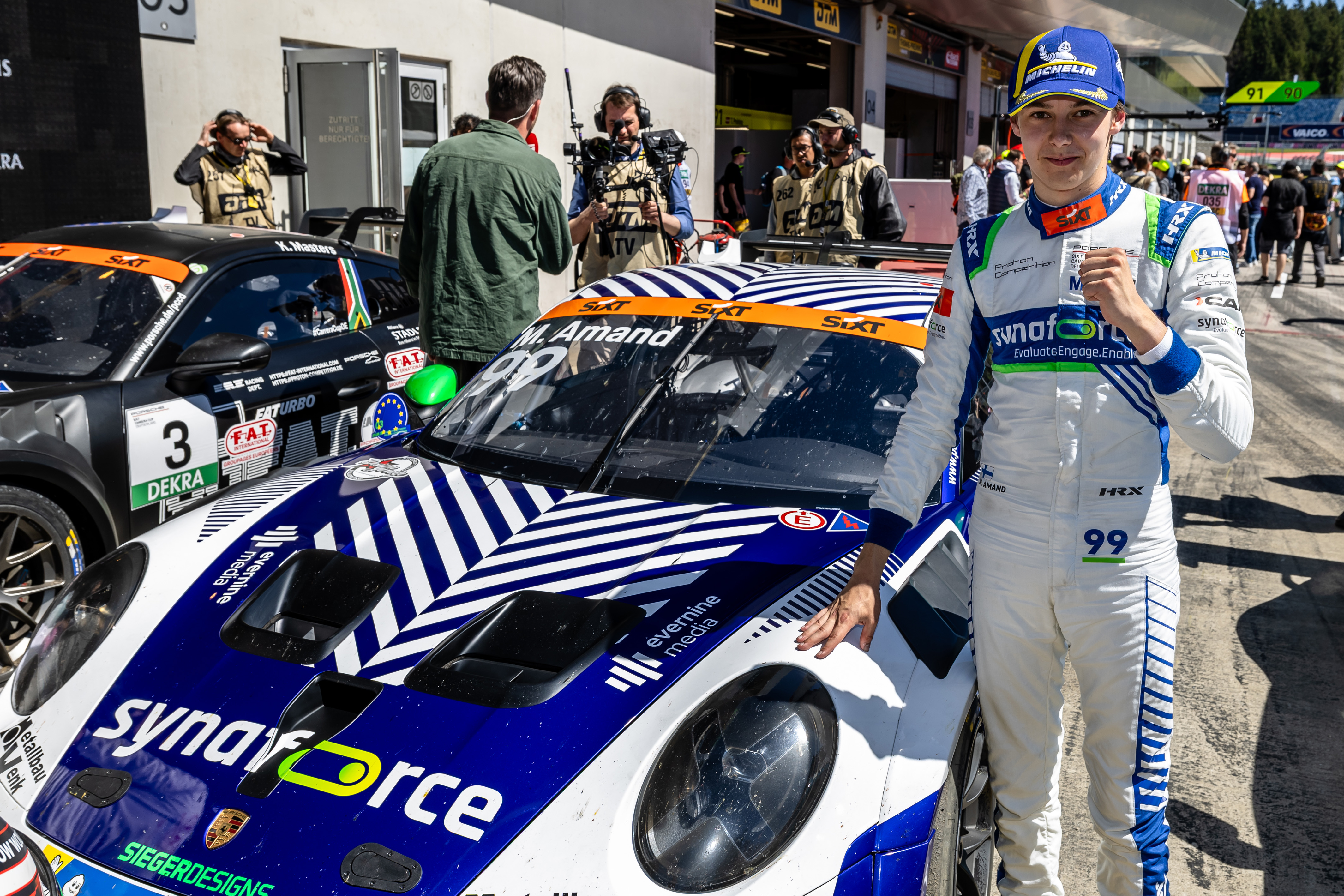 Racing driver M. Amand giving a thumbs up next to the white and blue Synaforce Porsche of Proton Competition with clearly visible Evernine Media logo, paddock at Red Bull Ring